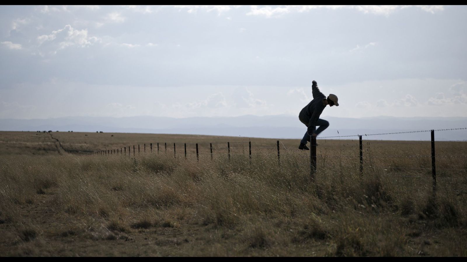 A scene from Australian film Alphabet Lane. A man wearing a cap jumps a barbed wire fence in a vast empty paddock.