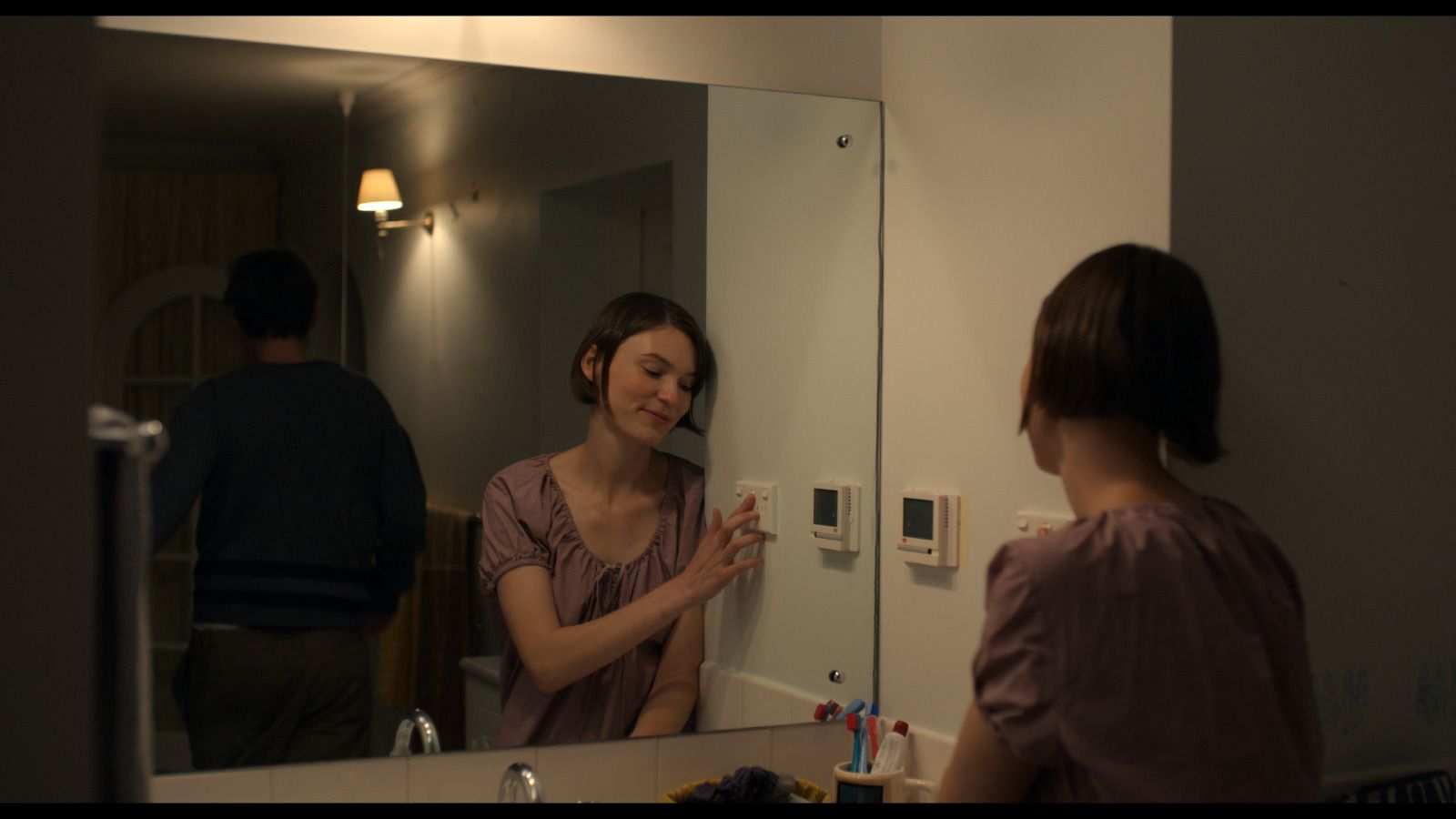 Anna (Tilda Cobham-Hervey) in a mauve blouse stands at a bathroom mirror, adjusting a wall panel, while Jack (Nicholas Denton) is visible in the background reflection.