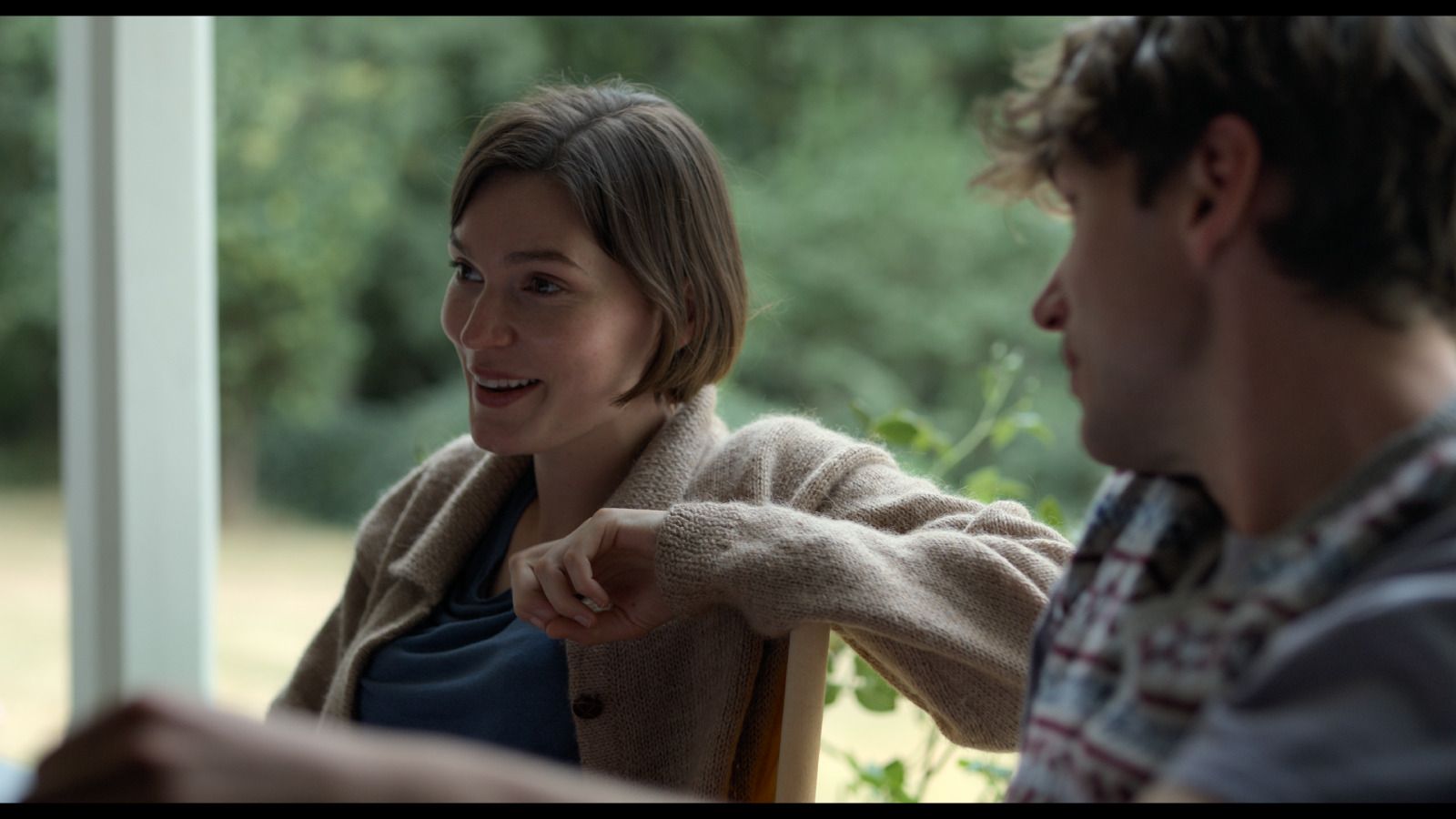 A scene from Alphabet Lane, where a woman (Anna) seated in a wooden chair laughs warmly toward a man (Jack) beside her, with a lush green landscape visible behind them.