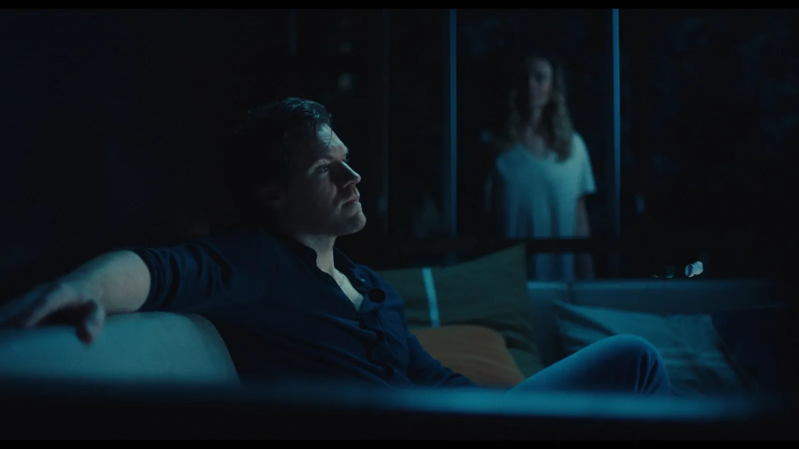 Jake (Alexander England) watches TV in the dark while his wife Sarah (Maeve Dermody) watches through the back door.
