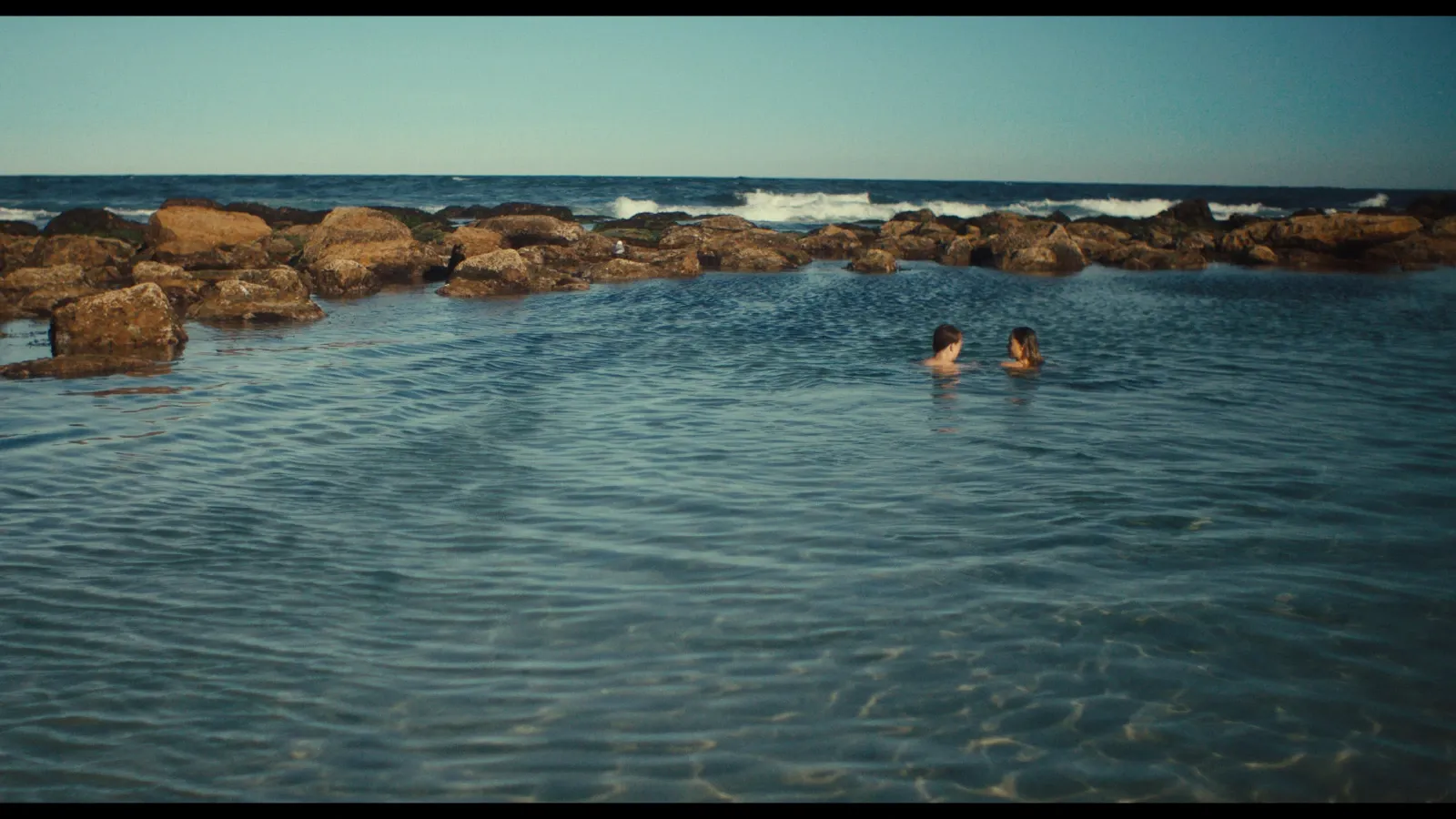 Sarah (Maeve Dermody) and Otis (Sonny McGee) swim in the rockpool of an Australian beach in Life Could Be A Dream — backlit by late afternoon sun.