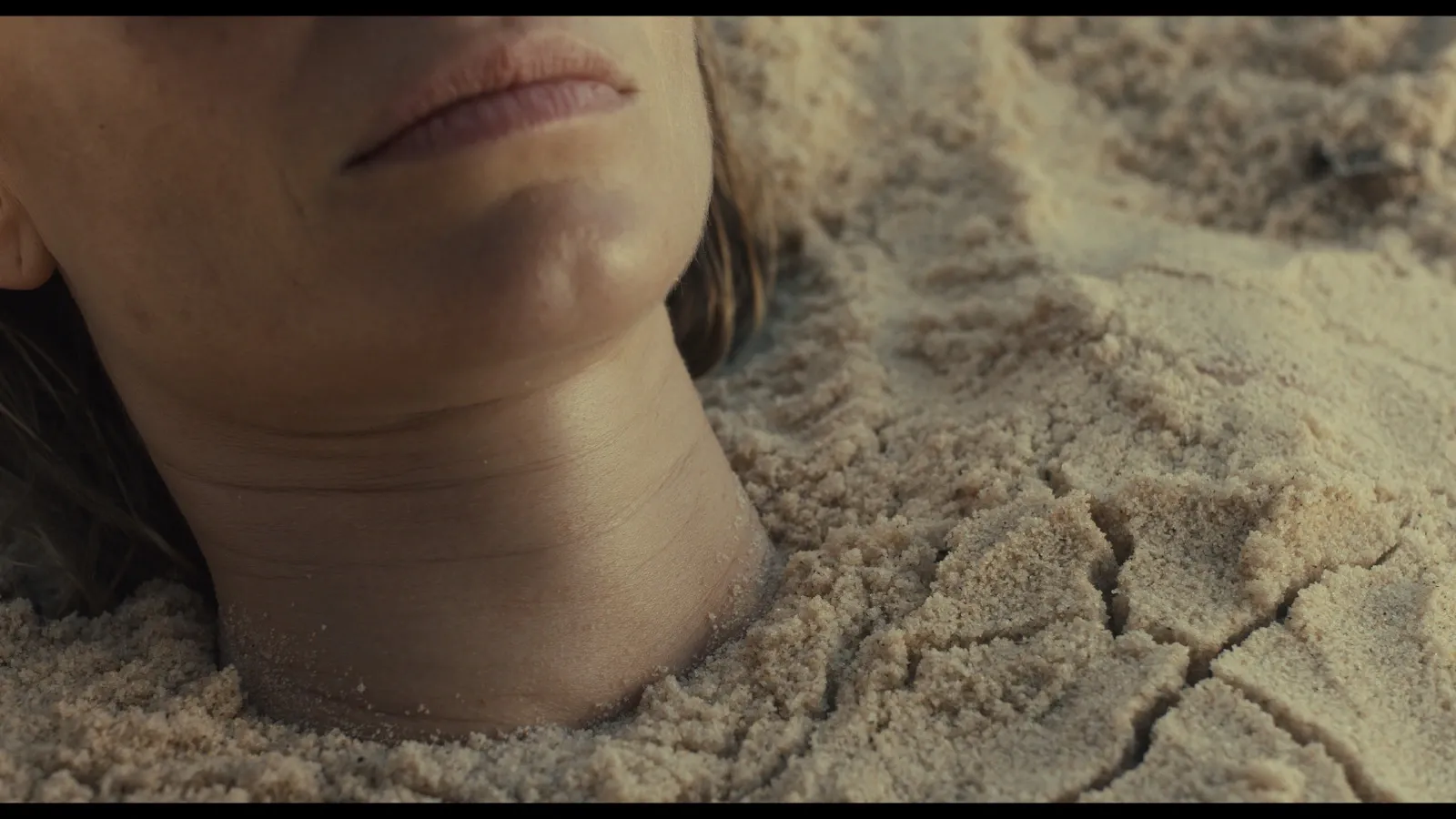 A close up of the neck and chin of Sarah (Maeve Dermody) buried at the beach, showing cracks in the sand
