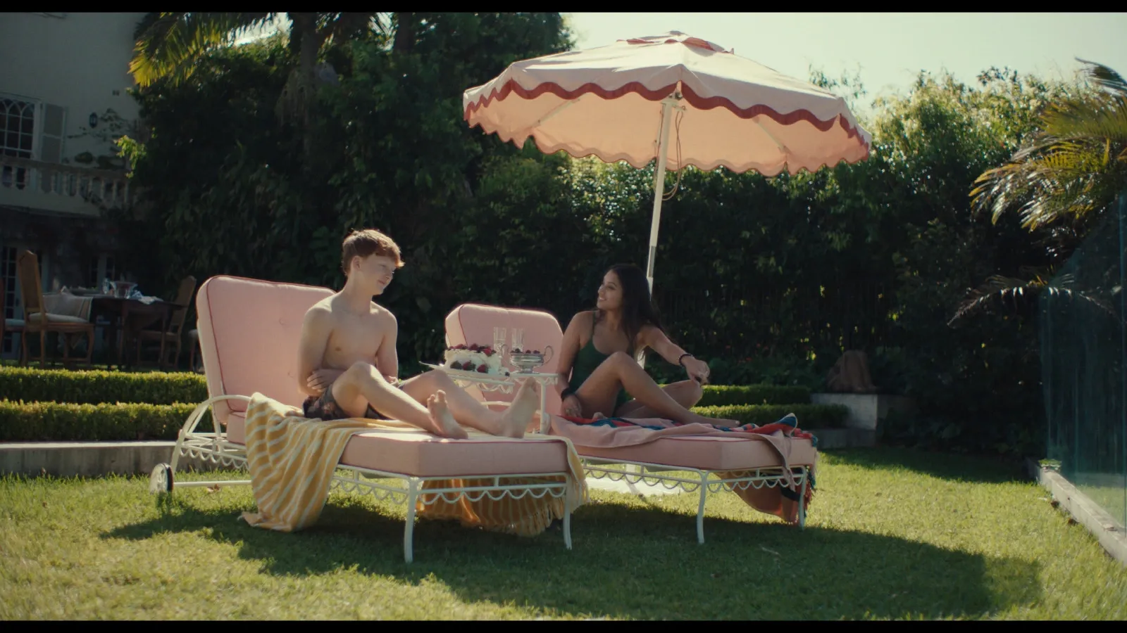Otis (Sonny McGee) relaxes on a pink plush deckchair by the pool next to Sati (Noam Sen-Gupta) a homemade pavlova 
